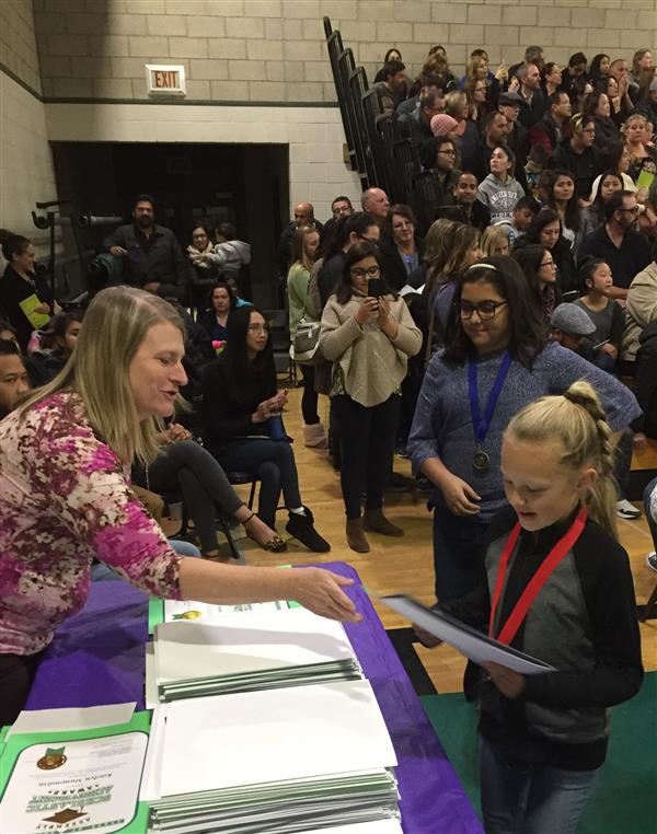 Teacher Tracy Buss hands a certificate to a student from a table. A crowd of people can be seen in the background.